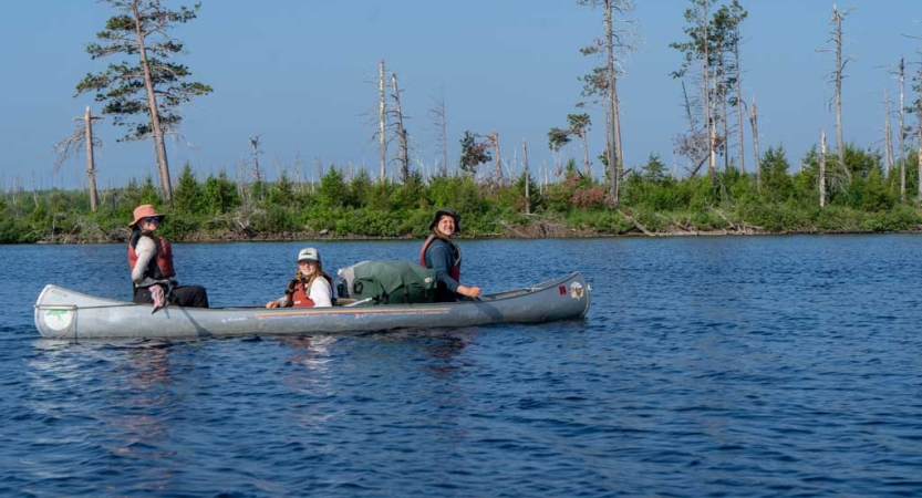 Three students navigate a canoe on calm blue water. There is a green shore in the background. 